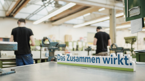 Auszubildende in der Werkstatt mit einem "Das Zusammen wirkt"-Schild im Vordergrund