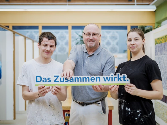 Zwei Malerei-Auszubildende halten mit ihrem Ausbilder ein "Das Zusammen wirkt"-Schild
